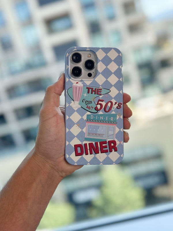 Hand holding a phone with Pela's The 50s Diner phone case, featuring a retro design with a blue and white checkered background. The case displays classic diner elements such as a milkshake, jukebox, and the text 