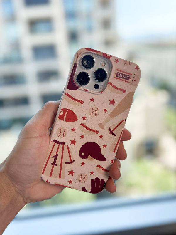 Hand holding a phone with a Pela Sunday Baseball Phone Case, featuring illustrations of baseball bats, balls, hotdogs, gloves, and jerseys in a playful, vintage style. The case has a beige background with maroon and tan accents, capturing the classic baseball game day vibe.