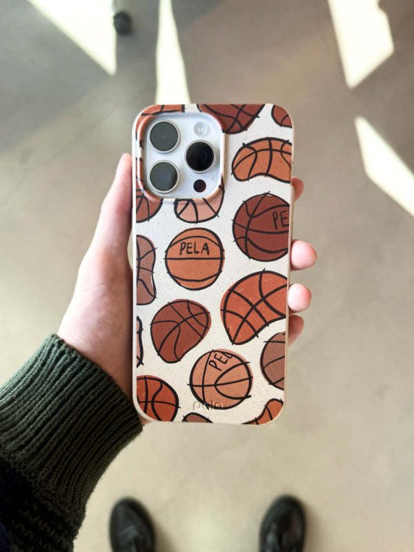 A hand holds the Pela Slam Dunk phone case, featuring a playful pattern of basketballs with “Pela” printed on some of the balls. The design is in warm shades of orange and brown on a light background, perfect for basketball fans who want to show off their passion.