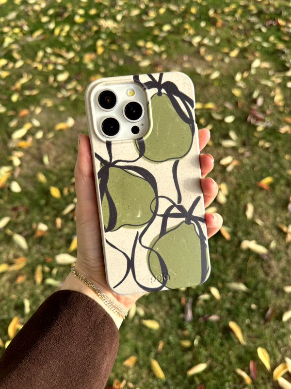 A hand is holding an iPhone featuring the Pela Sweet Pears phone case against a backdrop of grass sprinkled with fallen autumn leaves. The case showcases a bold design of large green pears outlined in black on a beige background. The earth-friendly case is paired with a brown jacket, complementing the fall-inspired scene and emphasizing the nature-themed design.