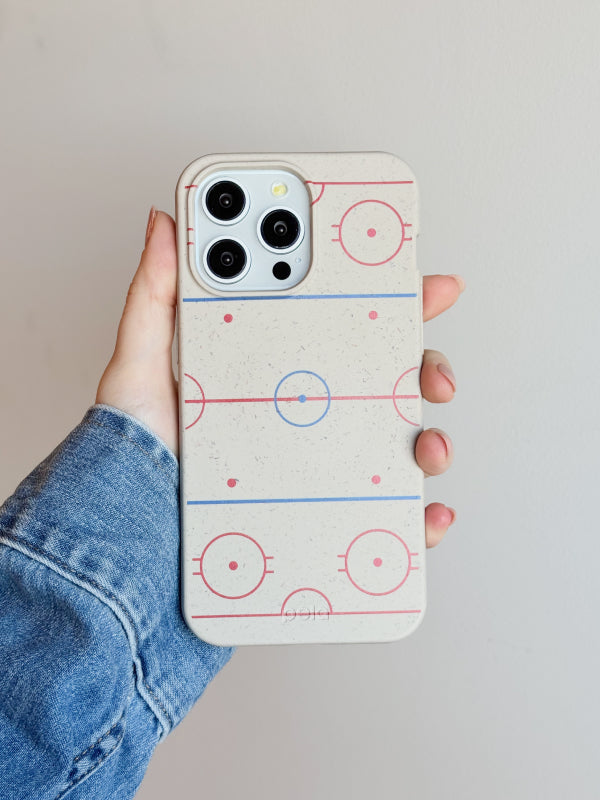 A hand holds an iPhone featuring the Pela Ice Rink Phone Case, which has a minimalist design of a hockey rink diagram in red and blue lines on a cream-colored background. The person’s denim jacket adds a casual, sporty vibe to the scene. The clean design and earth-friendly materials make this case a perfect choice for hockey enthusiasts.