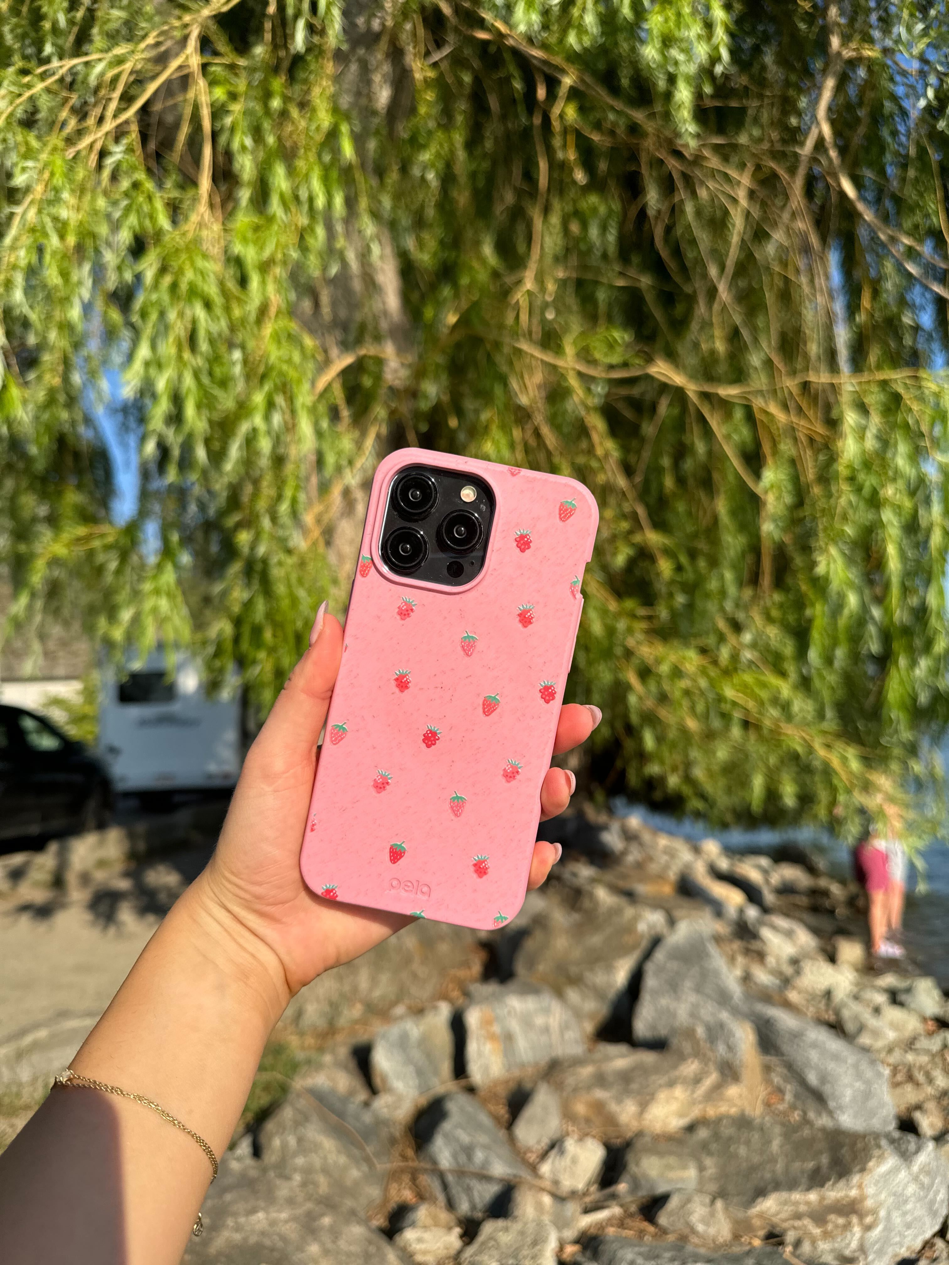 A hand holds Pela's Summer Berries Phone Case, which is pink and adorned with small strawberry illustrations. The background features a natural outdoor setting with greenery and rocks near the water.