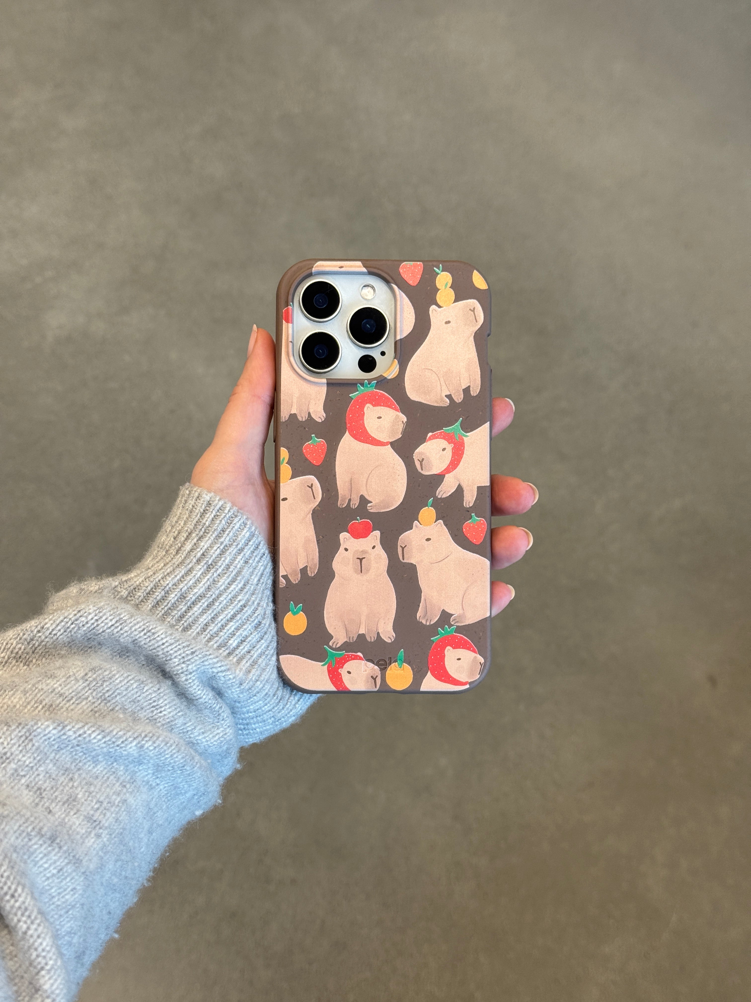 Hand in grey sweater holding iPhone with Pela Sweet Capybaras case featuring cream capybaras relaxing with red strawberries and orange fruits on chocolate brown background