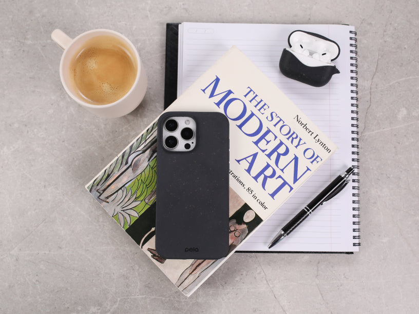  flat lay featuring the Pela Black Phone Case placed on top of a stack of books, including “The Story of Modern Art.” The scene is complemented by a notebook, pen, wireless earbuds, and a cup of coffee, creating a minimalist and stylish workspace aesthetic.