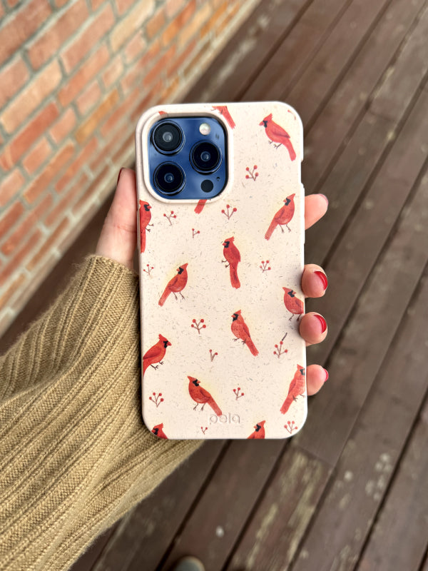 A hand holds an iPhone featuring the Pela Cardinals Phone Case, decorated with a charming pattern of red cardinals perched on a soft beige background. The person’s tan sweater and red nail polish complement the earthy, winter-inspired theme of the case. The background of a wooden deck and brick wall adds a warm, rustic touch to this earth-friendly, nature-inspired accessory.